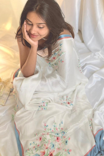 Woman wearing a white saree with floral patterns in a softly lit room.