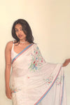 Woman wearing a white saree with floral embroidery against a plain background