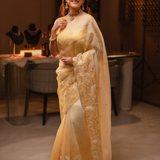 Woman in a yellow saree with intricate patterns standing in an indoor setting.