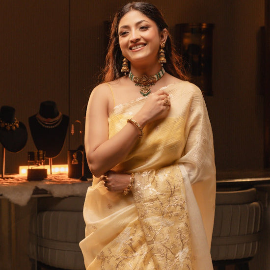 Woman in a yellow saree posing in a room with decorative items on tables.