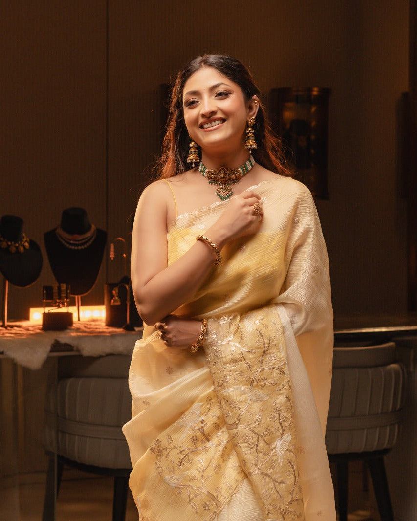 Woman in a yellow saree posing in a room with decorative items on tables.
