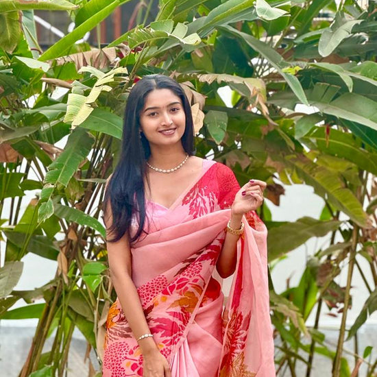 Woman in a pink saree with floral patterns standing among green plants.
