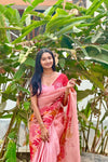 Woman in a pink saree with floral patterns standing among green plants.