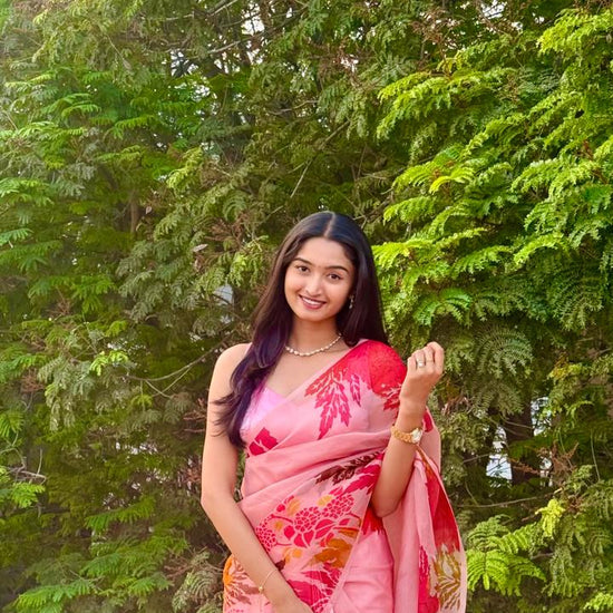 Woman in a pink saree with floral patterns standing in front of green trees.