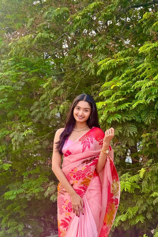 Woman in a pink saree with floral patterns standing in front of green trees.