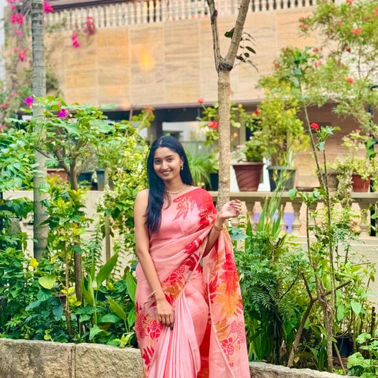 Woman in a pink saree standing in a garden with greenery and flowers around.